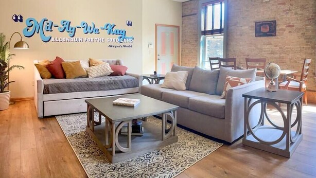 A cozy living room with a grey sofa, a coffee table, and a patterned rug. There's a brick wall and a ceiling fan. - Unit C- New Big 2 King Bedroom Condo by FiservForum/3rd St (Milwaukee)