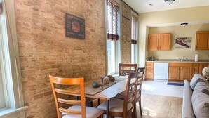 A dining area with a brick wall, wooden table, and chairs. There is a kitchen area with wooden cabinets and a countertop visible in the background. - Unit C- New Big 2 King Bedroom Condo by FiservForum/3rd St (Milwaukee)