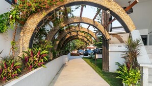Un pasillo cubierto con un arco tejido, rodeado de plantas en macetas y un sendero de hormigón.