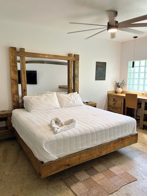 A spacious bedroom with a large bed, wooden headboard, a ceiling fan, a desk, and a window with natural light.