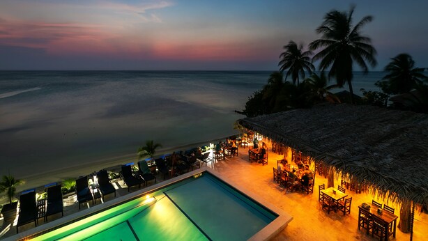 Zona junto a la piscina con sillones de descanso y una estructura de techo de paja, con vista a una playa al atardecer.