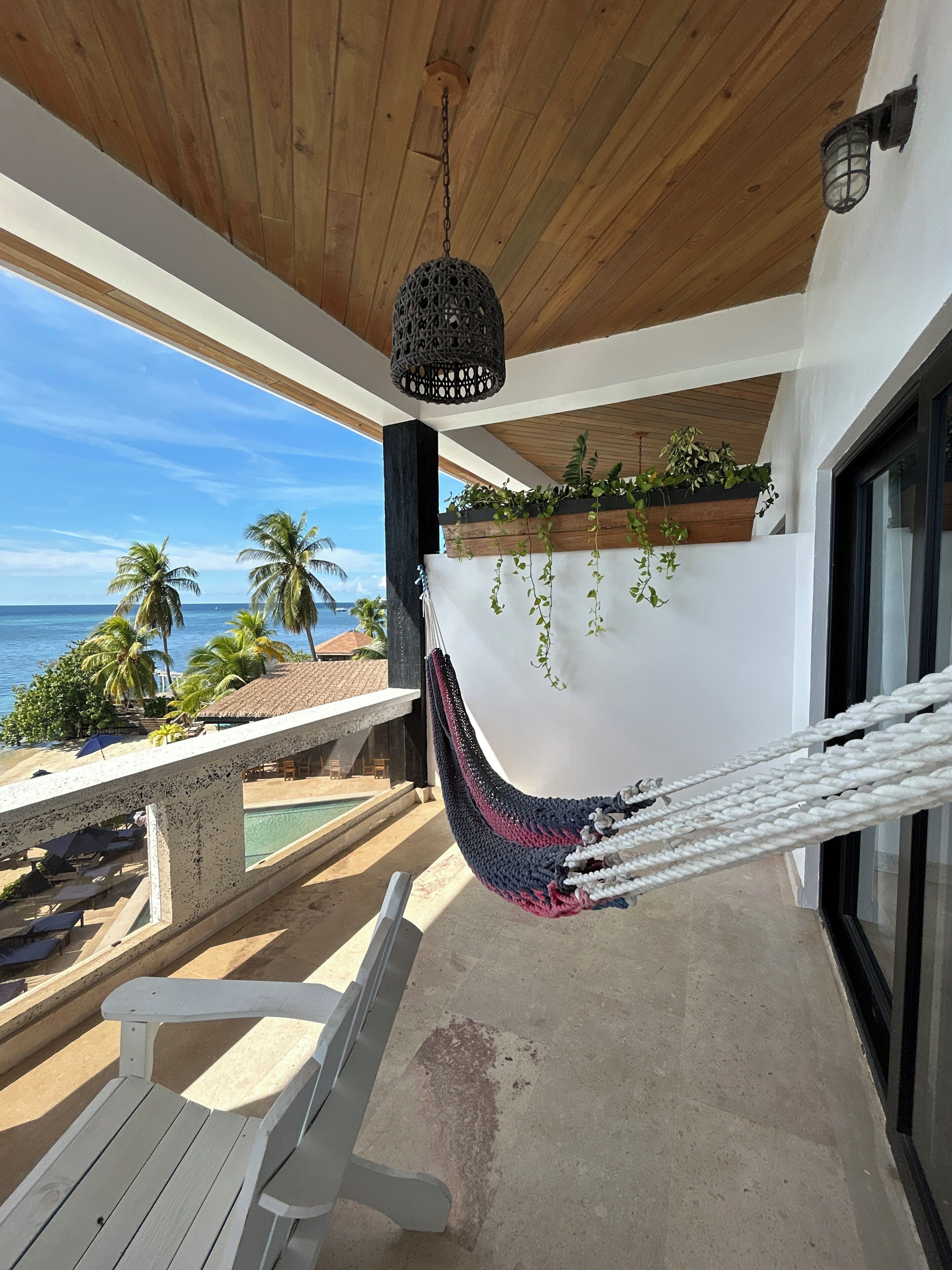 A hammock on a balcony overlooking a pool and beach with palm trees and ocean views.