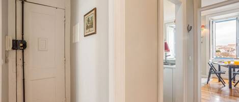 Un couloir avec une porte blanche, un tableau encadré au mur et une vue sur un coin repas avec une table et des chaises.