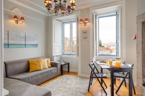A living room with a grey sofa, a dining table with chairs, and a chandelier. There are windows with blue frames and a view of buildings outside.