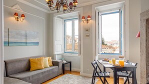 A living room with a grey sofa, a dining table with chairs, and a chandelier. There are windows with blue frames and a view of buildings outside.