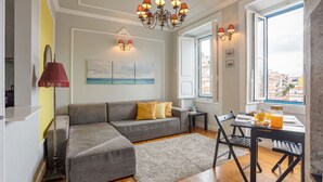A living room with a grey sofa, a coffee table, and a dining area with a table and chairs. There is a chandelier, a painting on the wall, and a window with a view of buildings.