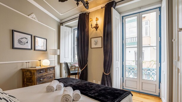 A hotel room with a bed, a desk, a chair, a dresser, and a balcony with curtains.