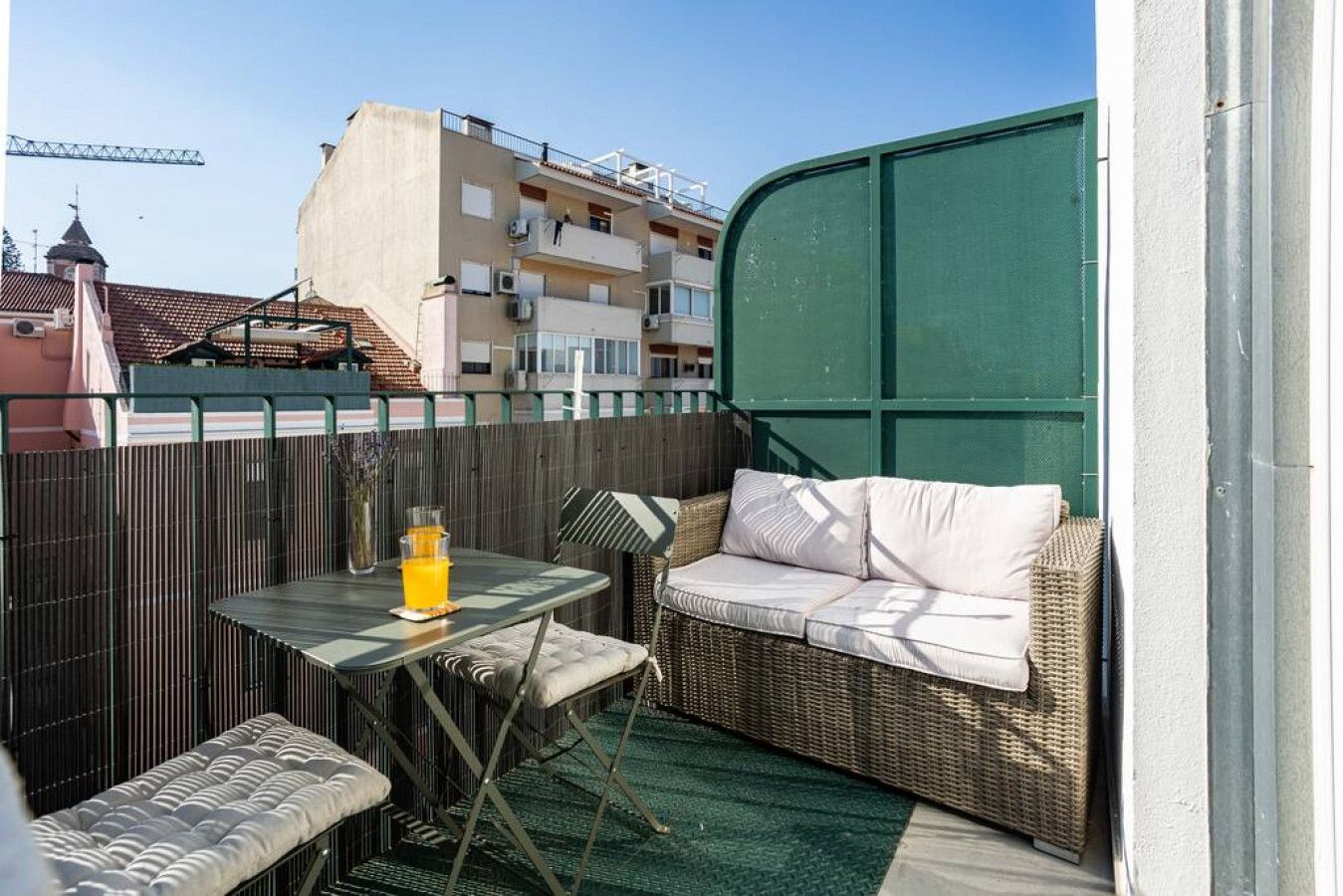A balcony with a table, chairs, and a sofa. There is a green wall and a fence in the background.