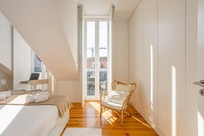 A bedroom with a bed, a laptop on a chair, a wicker chair, and a view of the outside through a large window.