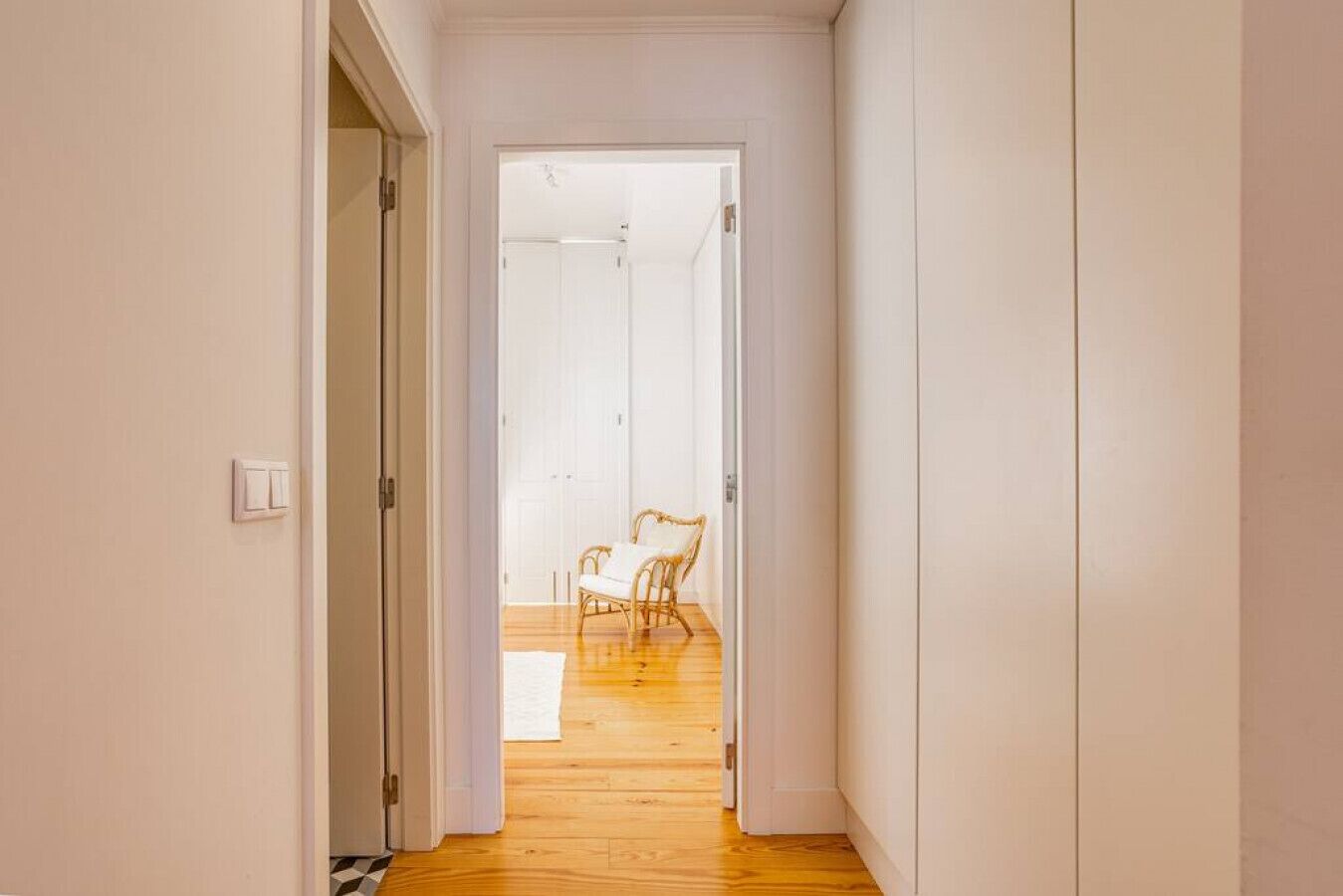 Um corredor estreito com piso de madeira que leva a um quarto com porta branca.