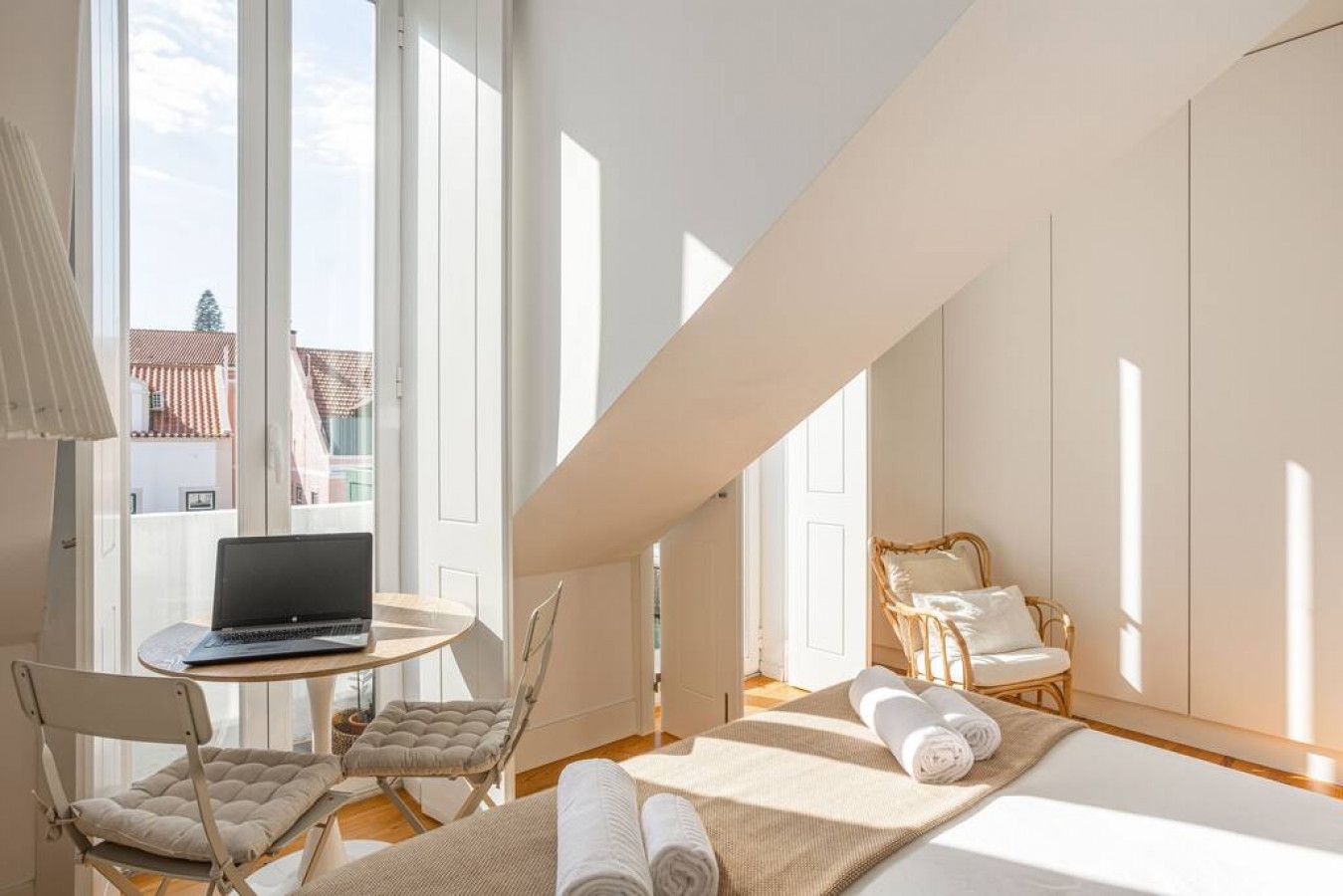 A bright room with a laptop on a small table, a cushioned chair, a wicker chair, and a bed with rolled towels.