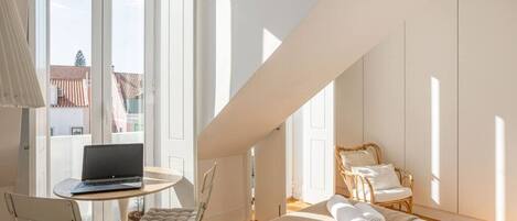 A bright room with a laptop on a small table, a cushioned chair, a wicker chair, and a bed with rolled towels.