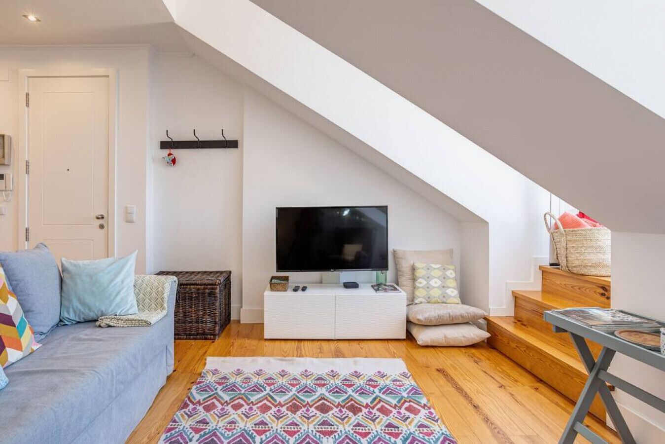 Ein gemütliches Lofts Apartment mit Fernseher auf weißem Schrank, einem gemusterten Teppich, einer Couch mit Kissen, einem Korbwäscheschrank und einer Treppe, die zu einem erhöhten Wohnbereich führt.