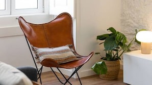 A room with a brown leather chair, a potted plant, a framed picture on the wall, and a white side table with a lamp. - Junqueira · Trendy T.M Apartament by the River (Lisboa)
