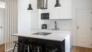 A modern kitchen with a central island, bar stools, pendant lights, and white cabinetry. - Junqueira · Trendy T.M Apartament by the River (Lisboa)