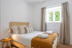 A bedroom with a wooden bed, white bedding, a bedside lamp, and a window with curtains.