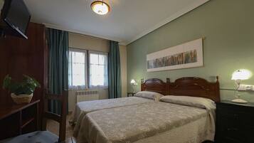 A bedroom with two beds, a television, a radiator, a window with curtains, a desk with a lamp, and a wall-mounted artwork.