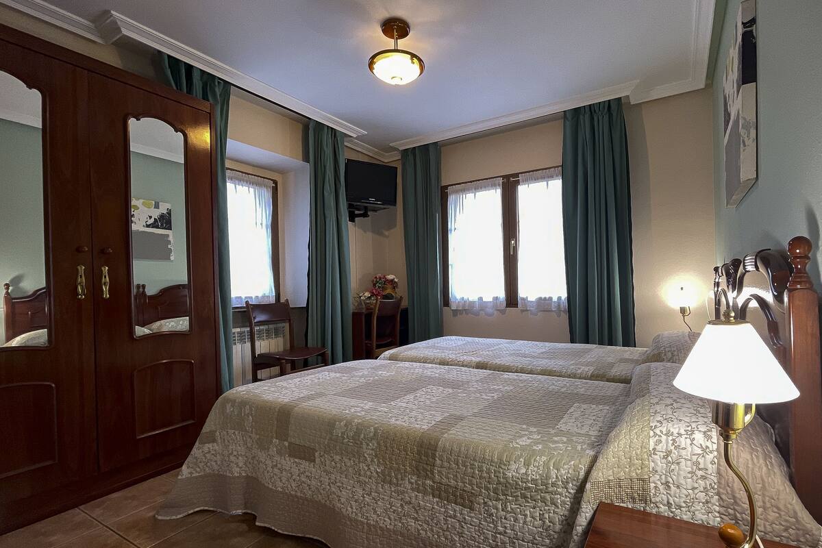 A hotel room with two beds, a wooden wardrobe, a desk with a chair, a TV, and a lamp.