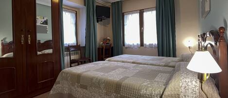 A hotel room with two beds, a wooden wardrobe, a desk with a chair, a TV, and a lamp.