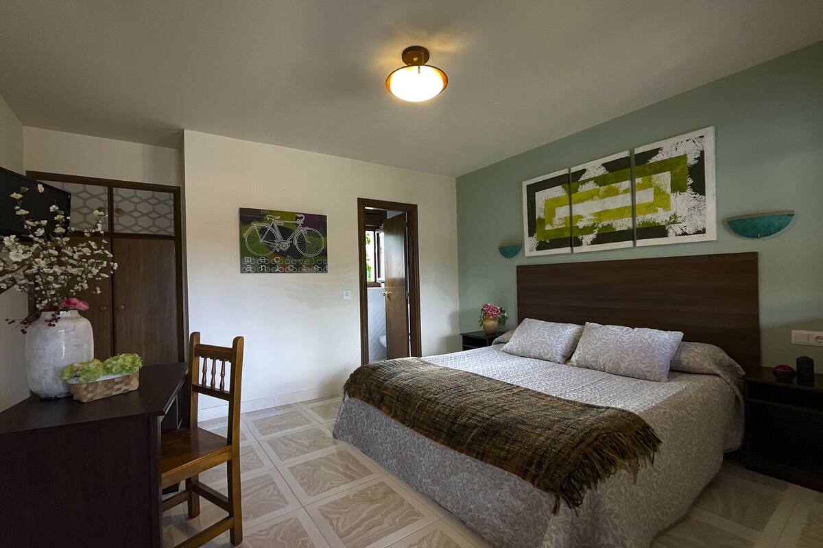 A bedroom with a bed, a desk with a chair, a vase with flowers, and a painting on the wall.