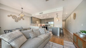 Un salon moderne avec un canapé gris, une table basse en bois et un coin repas comprenant une petite table et des chaises. On aperçoit la cuisine à l’arrière-plan, avec des appareils en acier inoxydable et un îlot central.