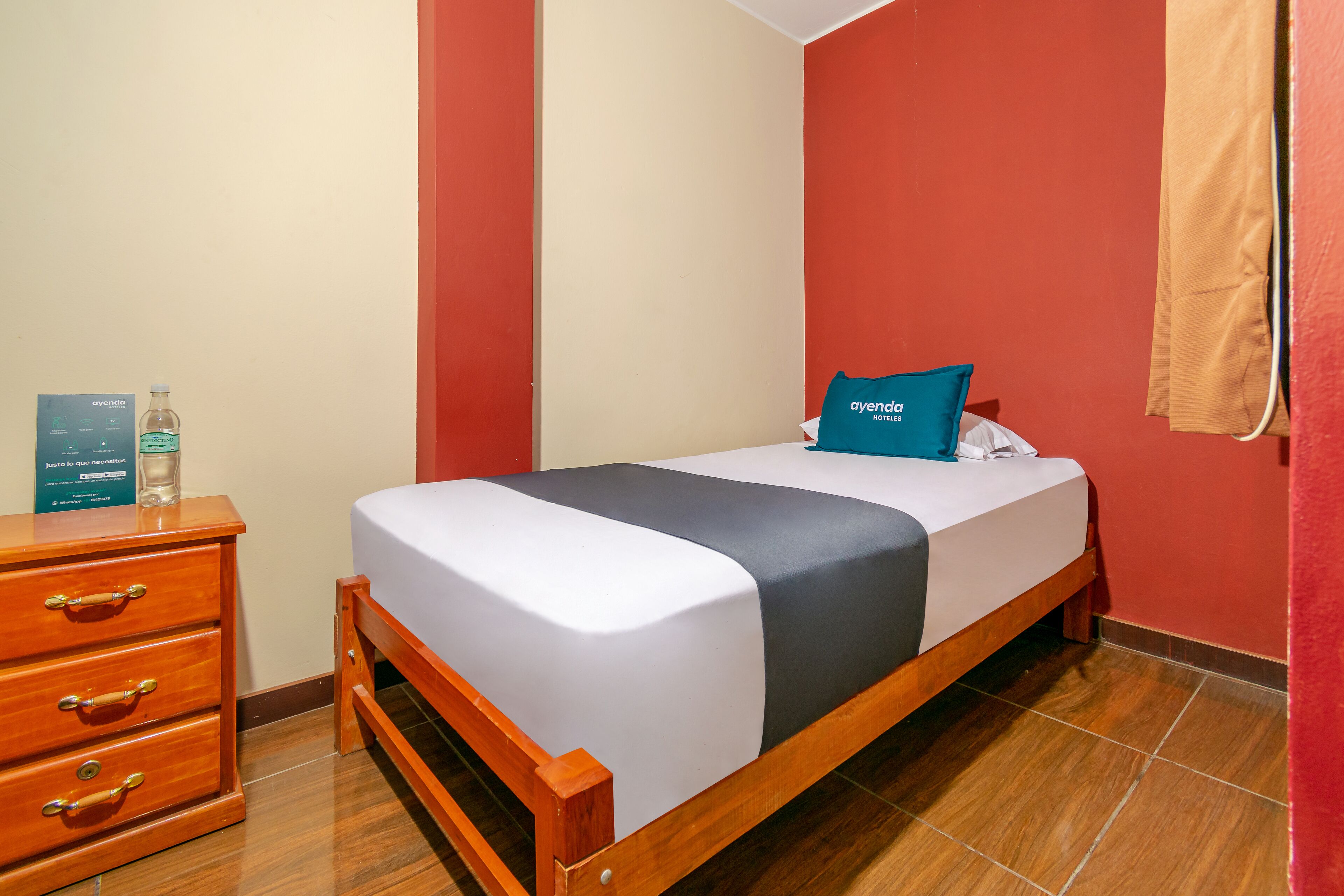 Una cama bien hecha con colcha gris y blanca, una mesita de noche de madera con cajones y una botella de agua encima.