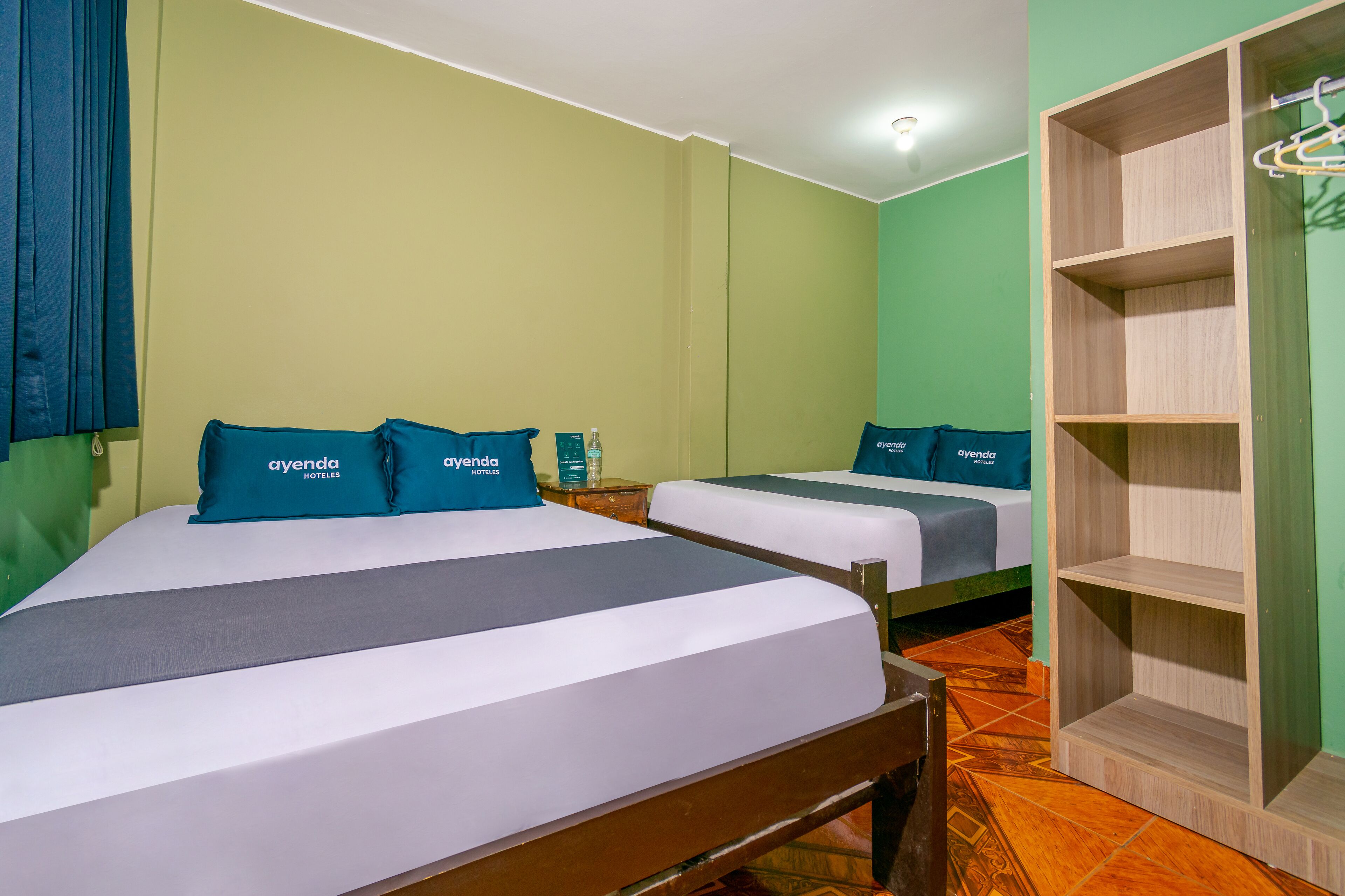 A hotel room with two beds, each with a blue pillow and a grey bedspread. There is a wooden wardrobe with a hanging rack on the right side.