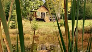 A wooden cabin with a deck, surrounded by a grassy field and a body of water.