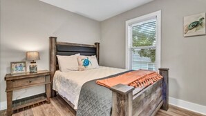 A bedroom with a wooden bed, a nightstand, a window with blinds, and a framed picture on the wall. - Large newly renovated family home. Sleeps 15. Walking distance to everything. (Oak Island)