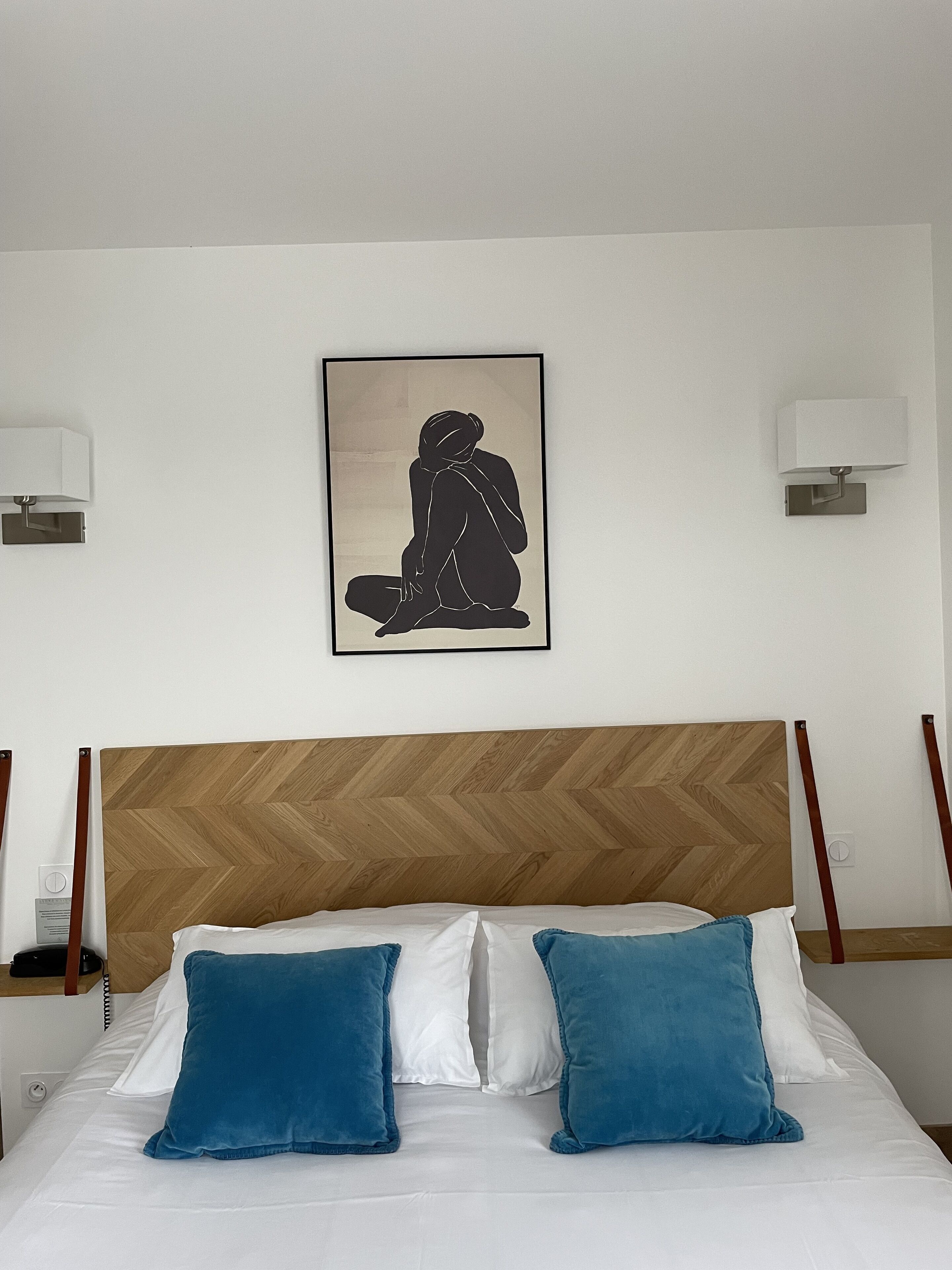Ein Hotelzimmer mit einem Holzboden-Kopfteil, einem Bett mit weißblauen Kissen und einem gerahmten Kunstwerk einer sitzenden Person an der Wand.