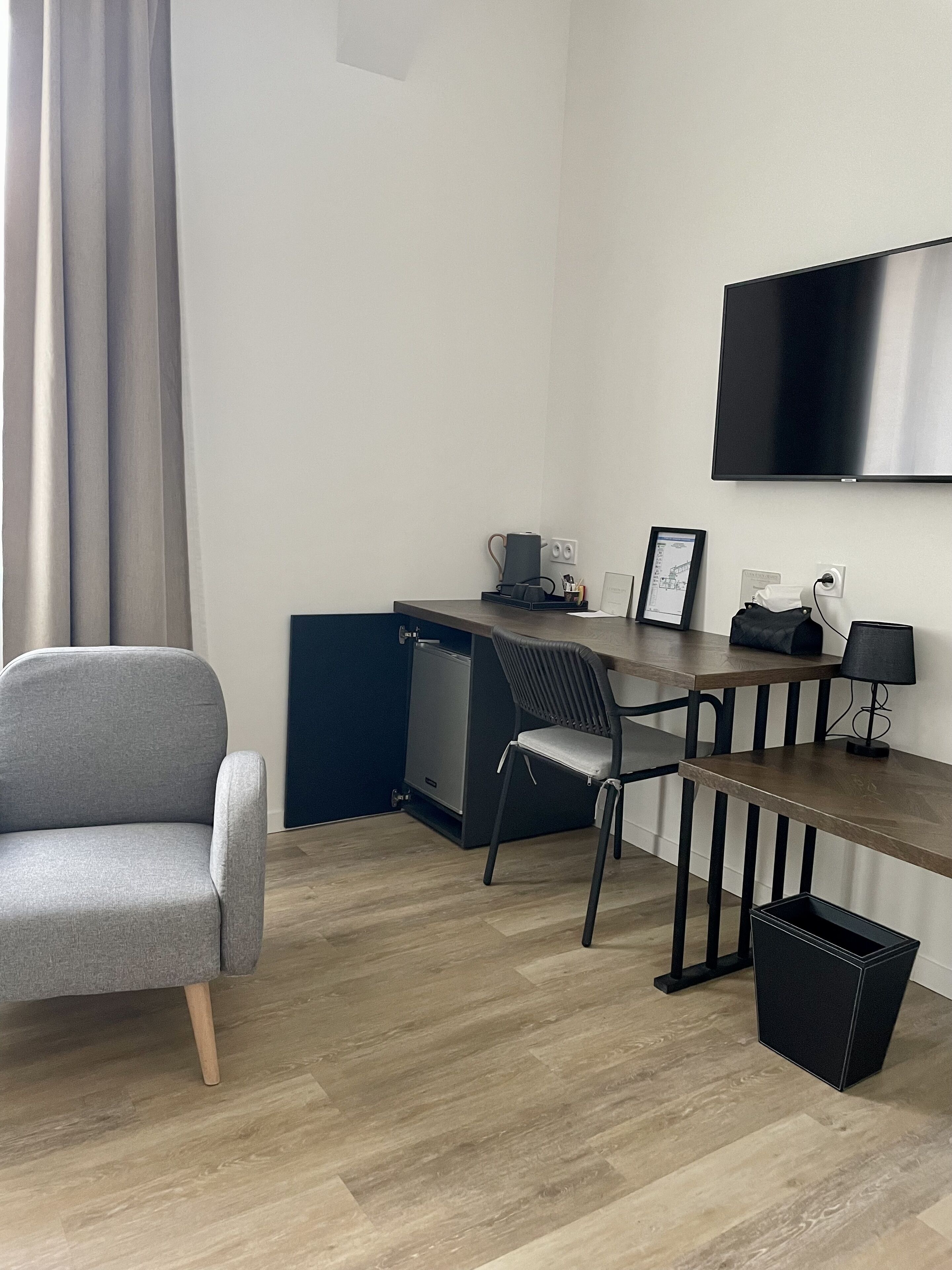 Una habitación moderna y minimalista con un sillón gris, un escritorio de madera, una silla negra, una pequeña kitchenette y un televisor de pantalla plana montado en la pared.