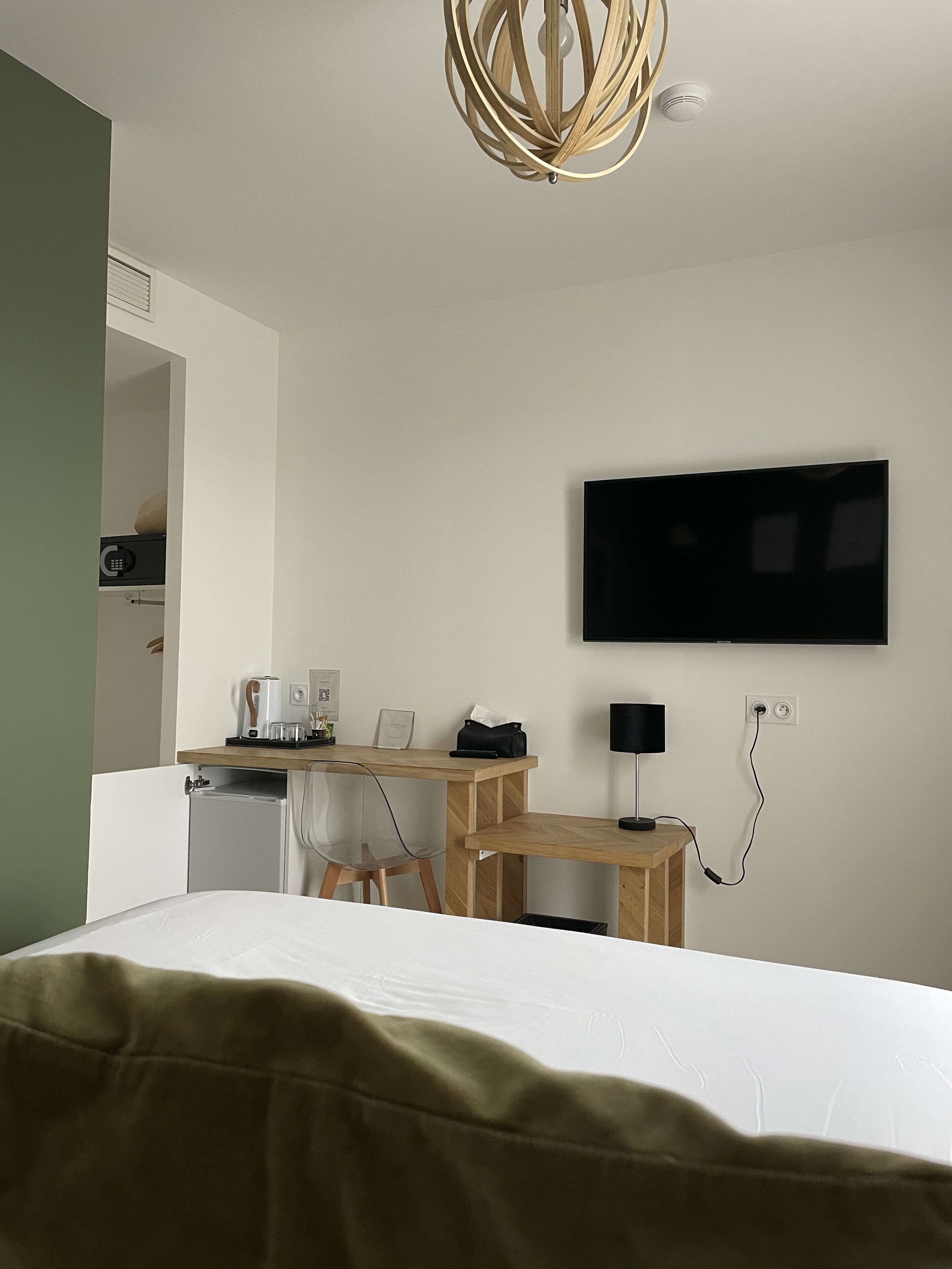 A modern hotel room with a bed, a desk with a computer, a television, and a small kitchenette.