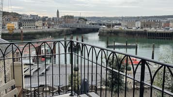 A riverside view with a wooden deck and metal railing, overlooking a town with buildings and a bridge.