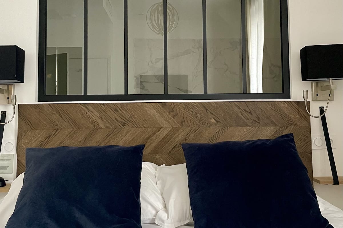 A hotel room with a large bed, two dark blue pillows, a wooden headboard, a large mirror, and two bedside lamps.