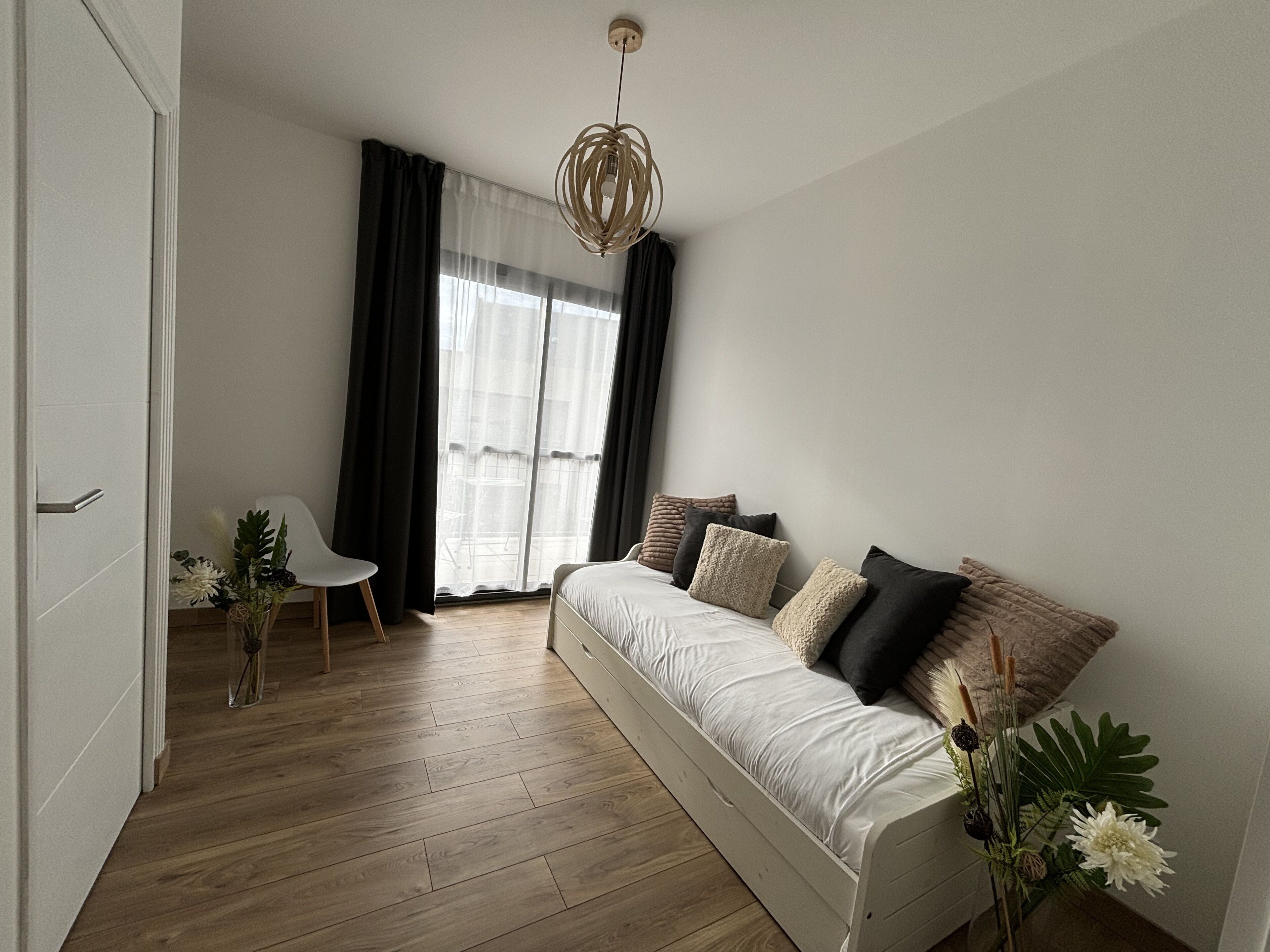 Ein modernes Schlafzimmer mit Holzboden, einem weißen Sofa mit vielen Kissen, einem Sessel und einem großen Fenster mit transparenten Vorhängen.