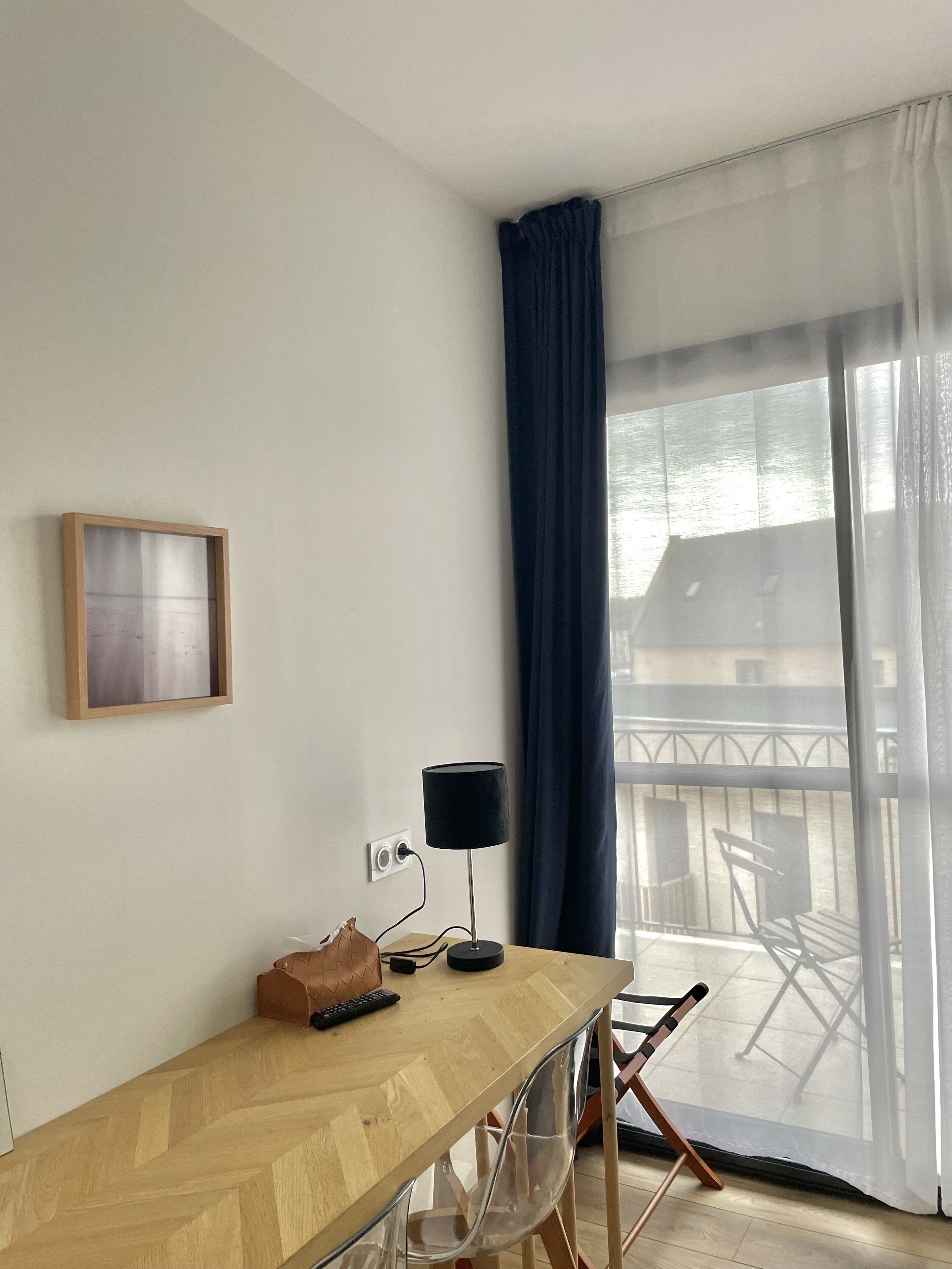 A room with a wooden desk, a black lamp, a framed picture, and a view through a large window with a blue curtain.