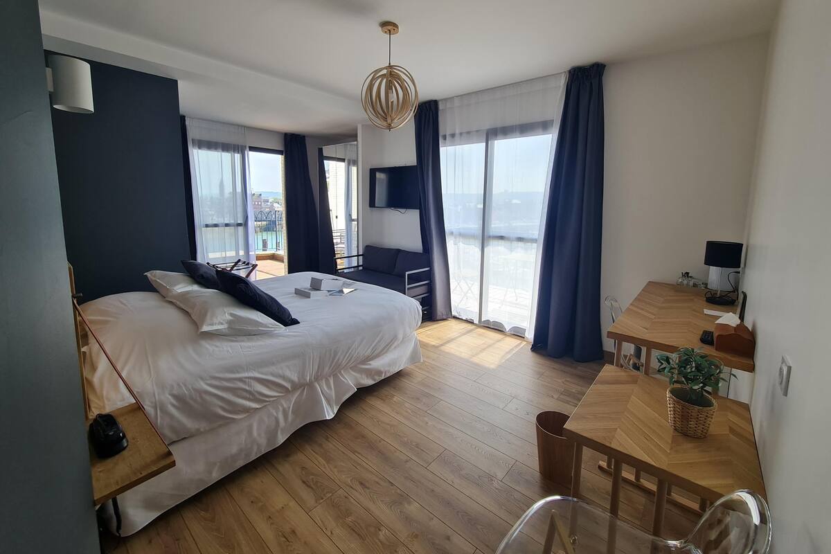 A modern hotel room with a large bed, a wooden desk, a chair, a TV, and a balcony with a view.