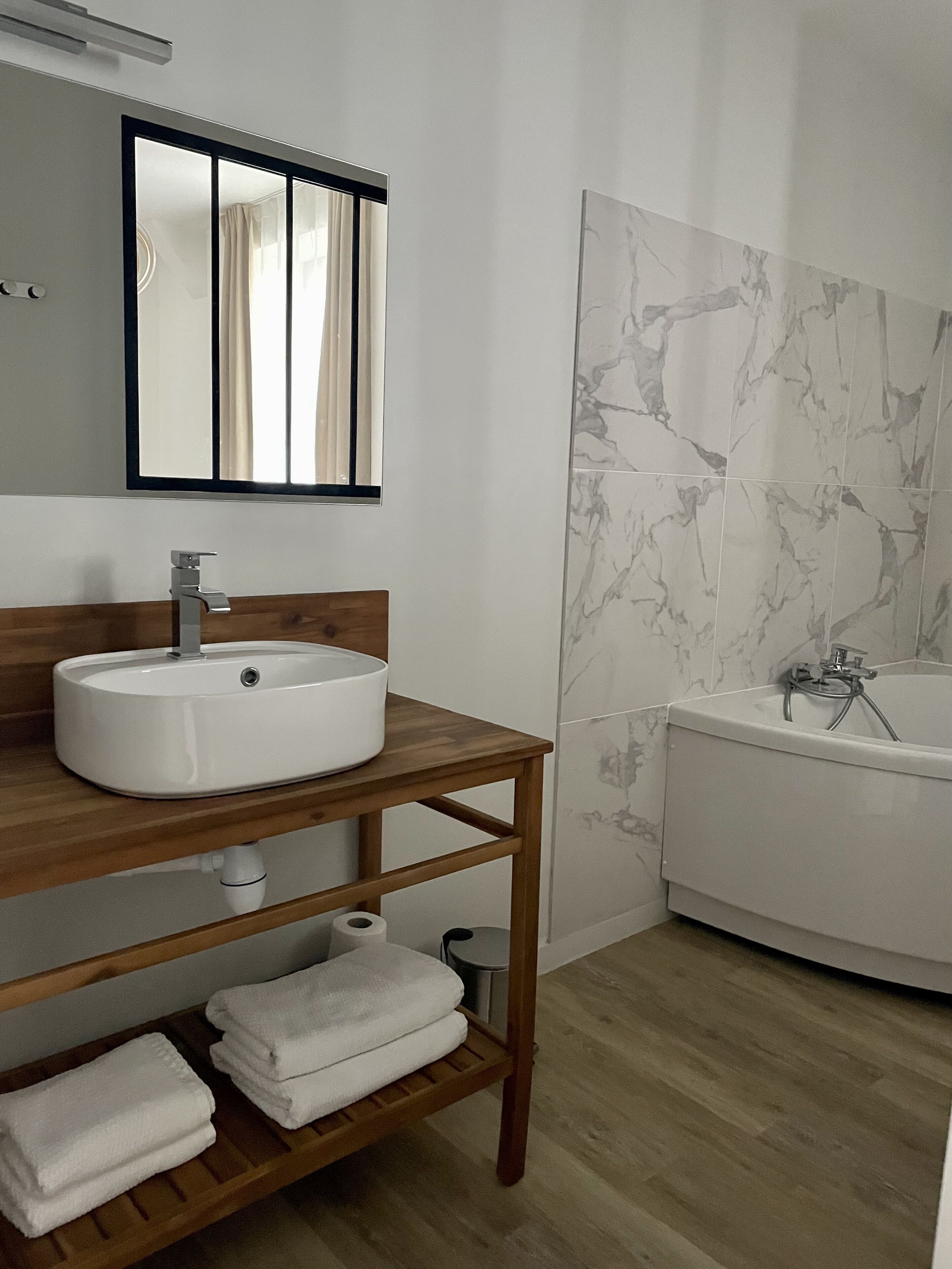 Une salle de bain avec un meuble-lavabo en bois, un lavabo blanc, un miroir, une fenêtre avec des rideaux, une baignoire recouverte de carreaux de marbre et un plancher en bois.