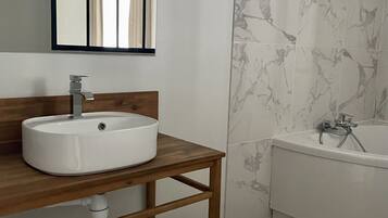 Une salle de bain avec un meuble-lavabo en bois, un lavabo blanc, un miroir, une fenêtre avec des rideaux, une baignoire recouverte de carreaux de marbre et un plancher en bois.