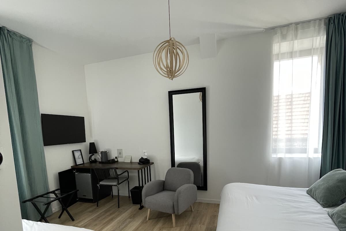 A modern hotel room with two beds, a desk with a chair, a small table, a mirror, and a hanging light fixture.