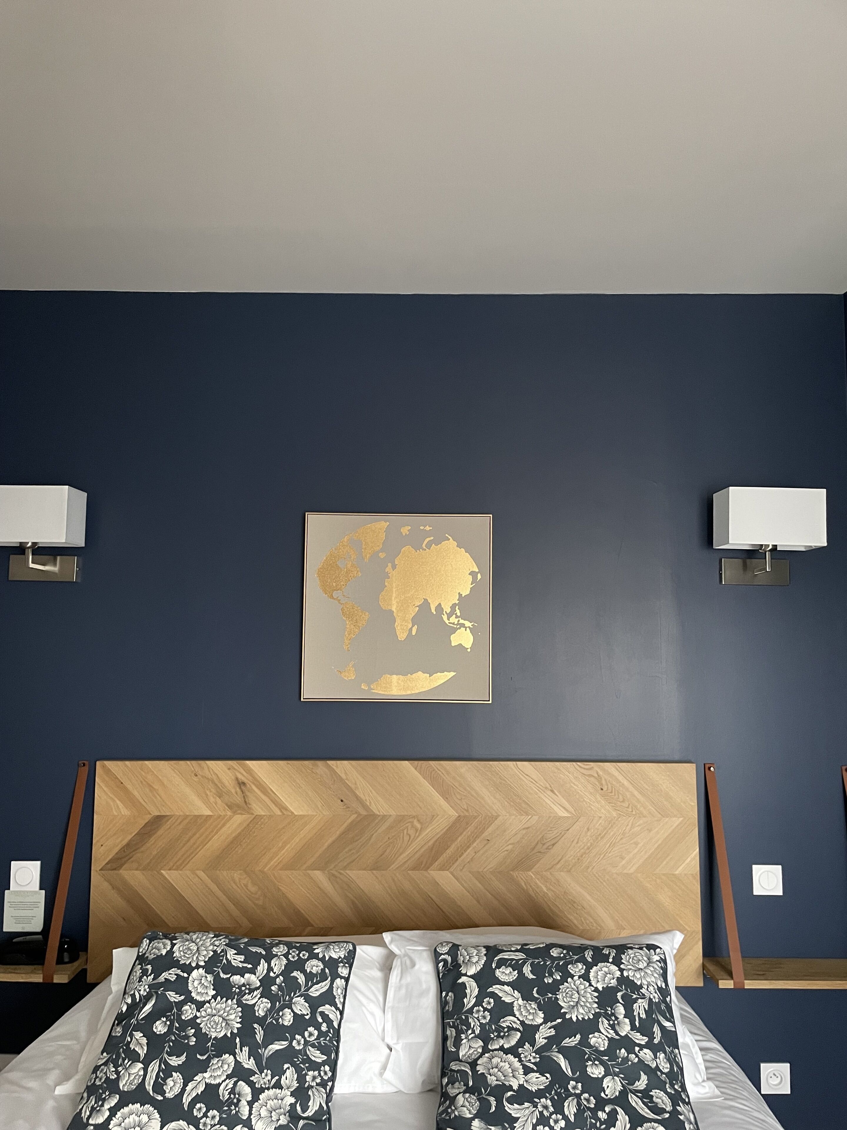 Habitación de hotel con cabecera de madera, cama con ropa de cama estampada con motivos florales, dos lámparas de noche y un mapa mundial enmarcado en la pared.
