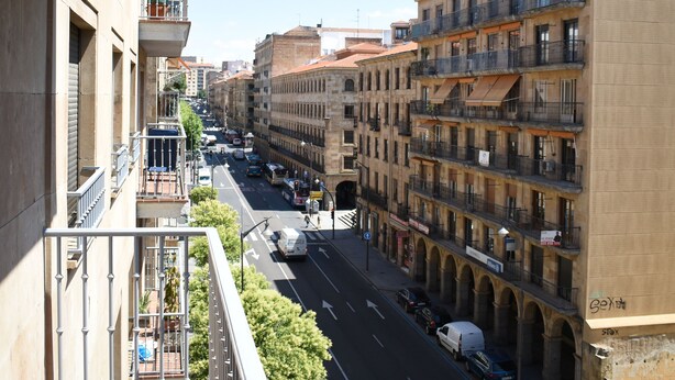 Un balcón con vista a una calle con edificios, un ciclista y coches estacionados.