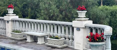 Zona junto a la piscina con barandilla de piedra, bancos y macetas con flores rojas.