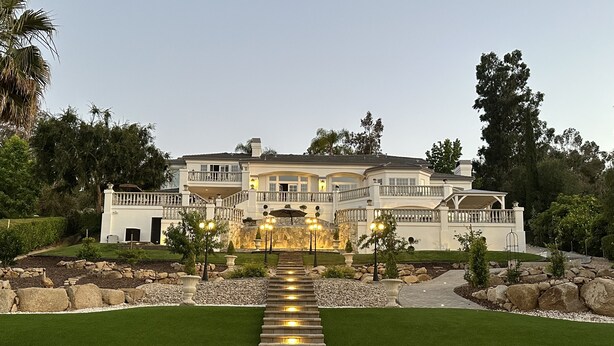 A large, multi-story house with a well-maintained lawn, a stone pathway leading to the entrance, and outdoor lighting.