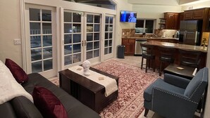 A spacious living room with a dark leather sofa, a blue armchair, and a coffee table. A large window with glass doors leads to a balcony. The kitchen is visible in the background with wooden cabinets and a stainless steel refrigerator.