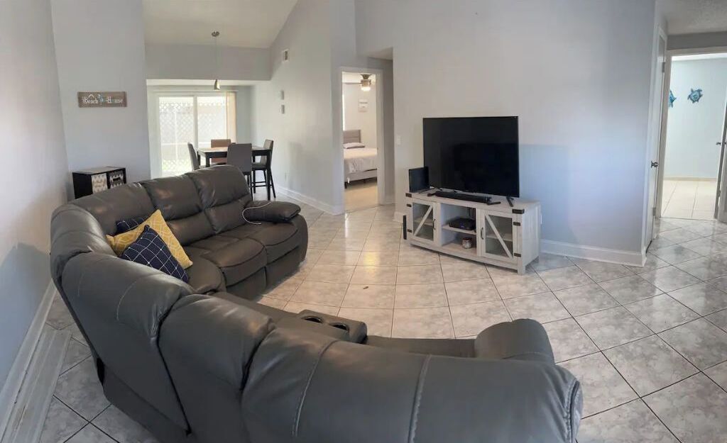 A modern living room with a leather sofa, a flat-screen TV on a white stand, and a dining area in the background.
