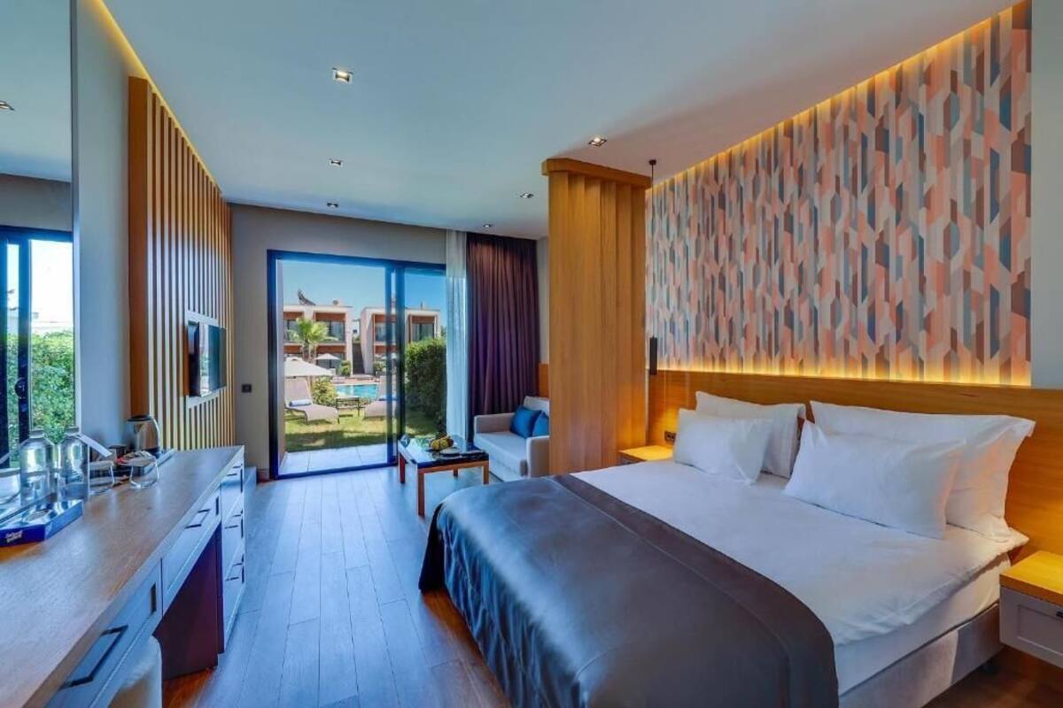 Quarto de hotel moderno com uma cama grande, uma escrivaninha, uma cadeira e vista para a piscina exterior e edifícios.