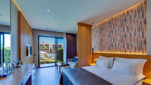 A modern hotel room with a large bed, a desk, a chair, and a view of the outdoor pool and buildings.