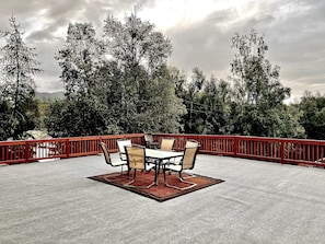 En udendørs terrasse med rødt gelænder, et bord med fire stole og udsigt til tætte træer og bjerge i baggrunden.
