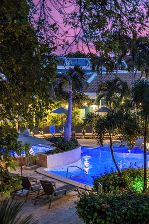 A pool area with lounge chairs and umbrellas, surrounded by palm trees and a building with a tiled roof. - Kura Botanica Hotel (Willemstad)